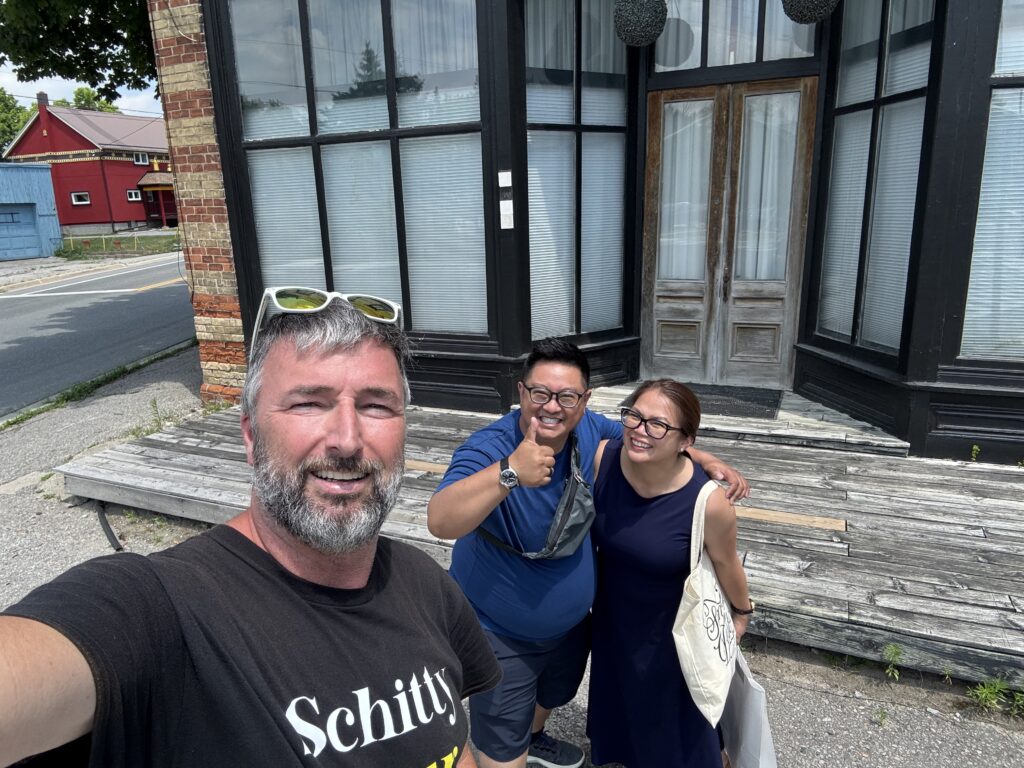 Two men, one giving a thumbs up, and a woman carrying a white tote bag taking a photo in front of the Cafe Tropical location.