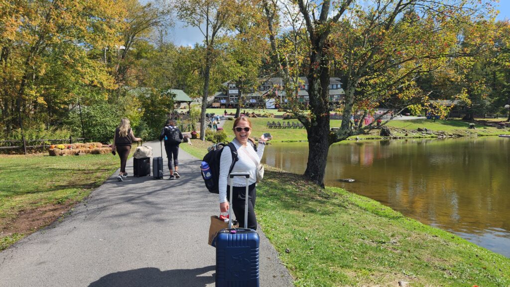 Woman in long sleeves smiling with luggage and backpack surrounded by trees and a lake in front of her