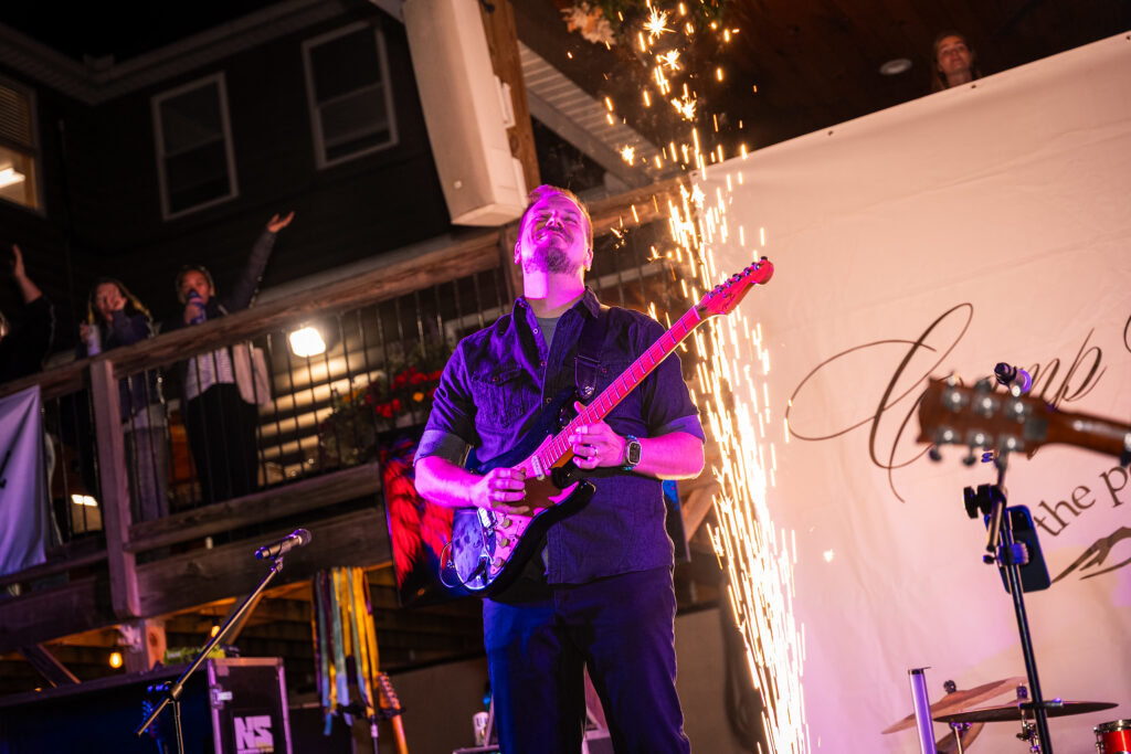 man playing an electric guitar with fireworks in teh background and 2 women waving in the background