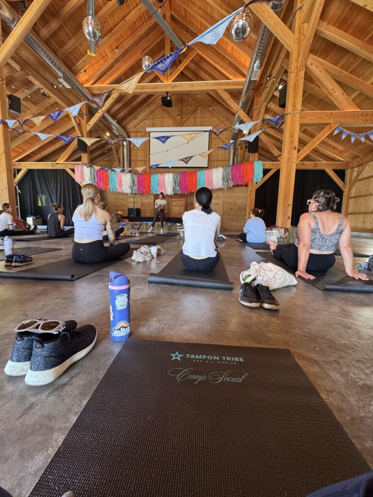 women sitting on yoga mat. mat says tampon tribe, camp social. purple owala bottle on display.