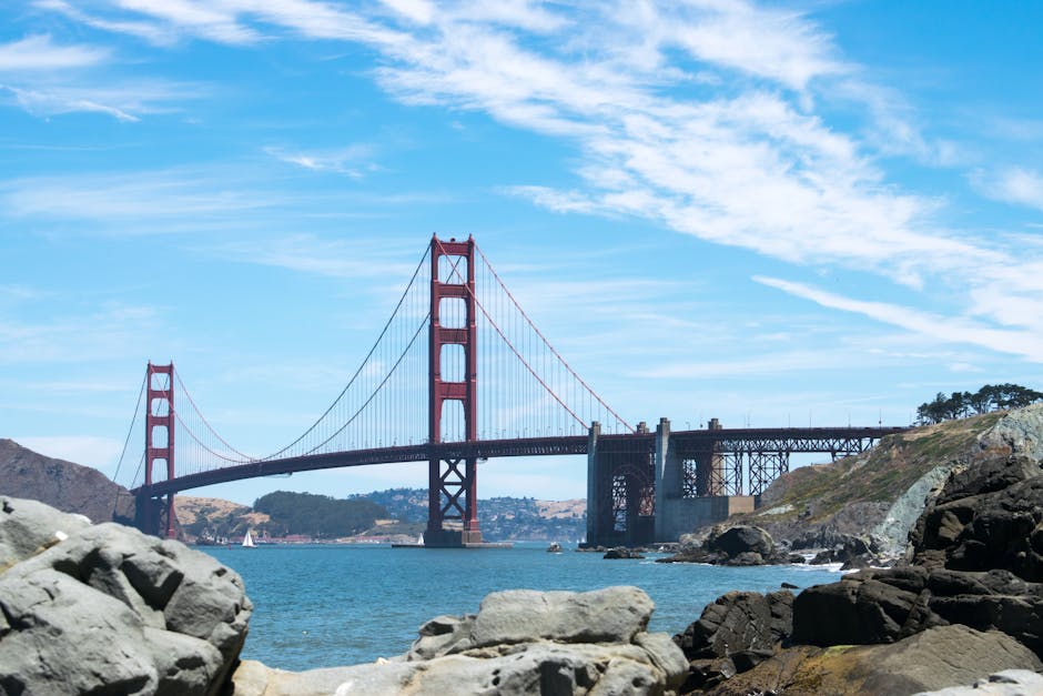 Panoramic view of the iconic Golden Gate Bridge spanning a bright blue ocean with scenic rocks in the foreground.